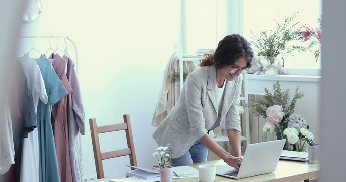 Happy young business woman designer using laptop taking online shop orders stand in atelier office. Smiling fashion retail store owner communicate with customers on website manage e business concept.