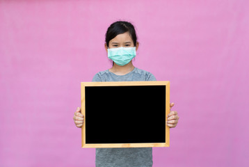 Little asian girl in a protective medical mask holding black board isolated on pink background. Protect from Coronavirus or Covid-19 epidemic.