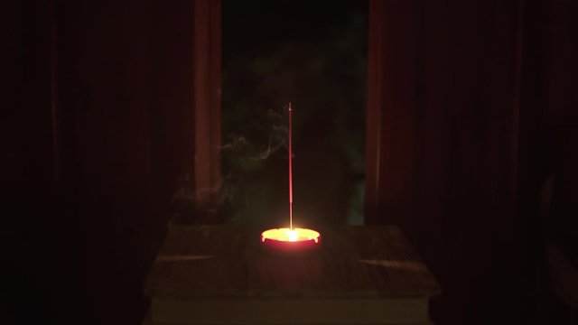 Incense stick and candle are burning and smoke on small table in dark room on background of open doors at night. Candle light. Spirituality magic ritual. Aromatherapy. Candle flame meditation relax