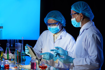 Laboratory technicians in medical masks discussing research details on tablet computer before...