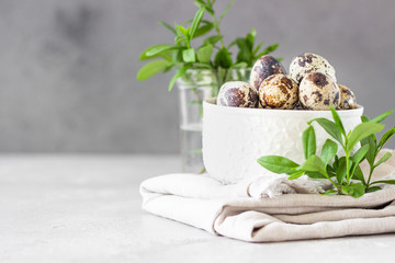 White ceramic bowl with organic quail eggs on linen napkin and green leaves, light grey stone background. Selective focus.
