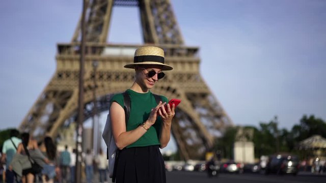 Slow motion of happy female tourist laughing and chatting in social networks on smartphone enjoying trip in France.Young woman making photo on mobile phone standing on street with Eiffel Towel
