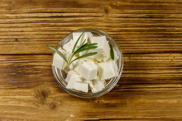 Feta cheese cubes with rosemary in glass bowl on a wooden table. Top view
