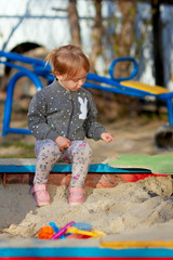 Adorable little girl sitting on sandbox on the playground on sunny spring day