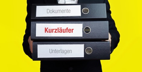 Kurzläufer. Mann im Anzug trägt einen Stapel Aktenordner. Hintergrund gelb. Ein Ordner hat eine...