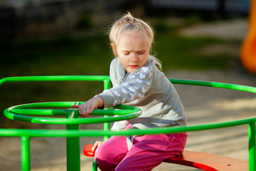 Pretty toddler blond girl playing on carousel on the playground on sunny spring day