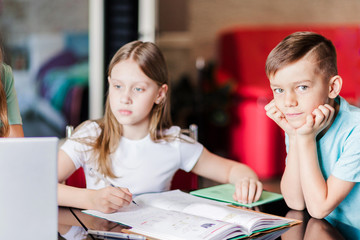 Girl with mom doing school lessons at home. Boy, little brother stands nearby and looks at the camera