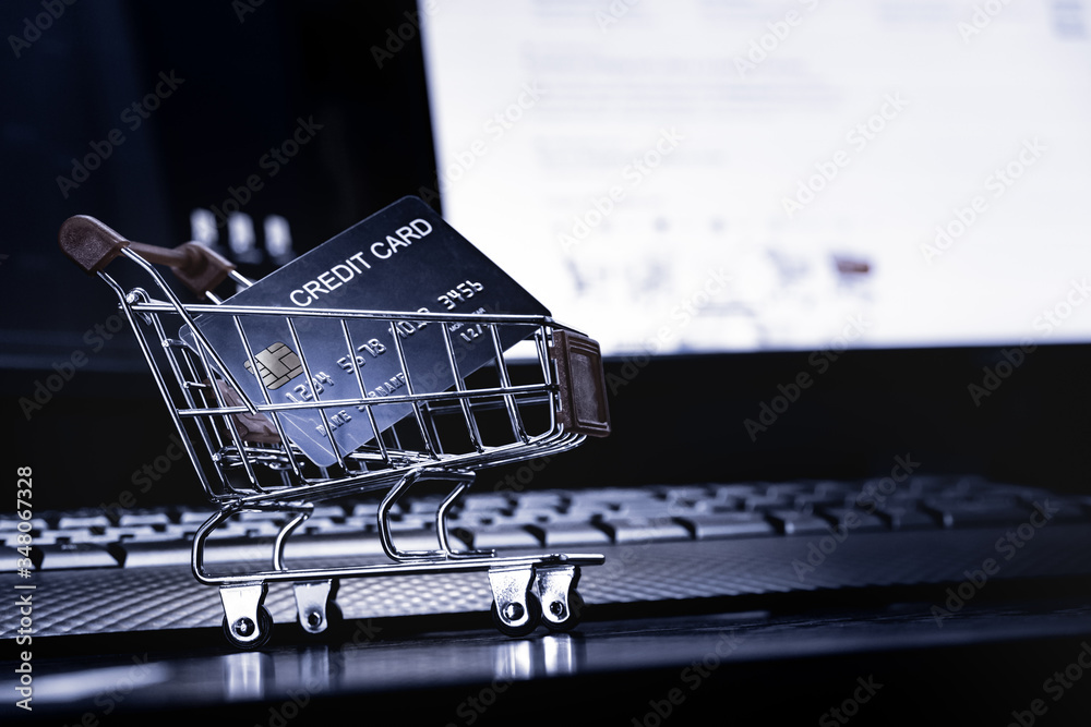 Poster close up Shallow focus Credit Card in mini shopping card with Computer keyboard and E-commerce website on monitor background. Background for E-commerce and online shopping concept.
