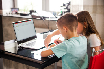 Fototapeta premium Boy and girl, brother and sister study at home. On the table are a laptop, books, notebooks, pencils and pens. Sister helps brother do homework