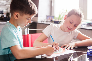 Boy and girl, brother and sister draw a rainbow with crayons together, sitting at a table