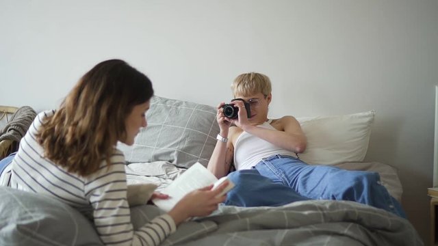 Young lesbian couple takes pictures and reads while lying on bed in bedroom. Spbd Homosexual american woman is reading books and posing with smile in front of camera in modern interior. Two lgbt girls