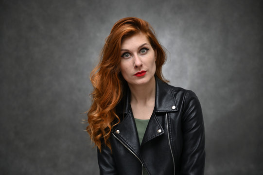 Studio photo of a young caucasian woman with long red hair in a black jacket. Pretty model actress posing with different emotions on a gray background in the studio.