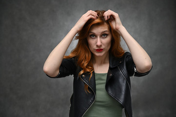 Photo of a young caucasian woman with excellent makeup with long red hair in a black jacket. Caucasian model actress posing with different emotions on a gray background in the studio.