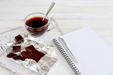 A cup of coffee, chocolate, a sheet of paper and a pen on a white wooden surface. Background for work at home in quarantine. The difficulty of adapting work at home.