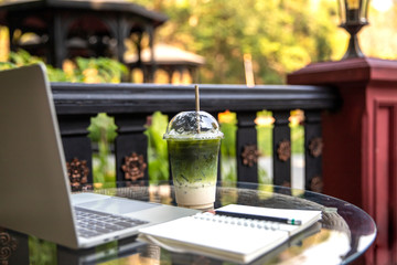 Matcha Milk , Milk Green tea latte with laptop and note book on glass table  outdoors on nature  background