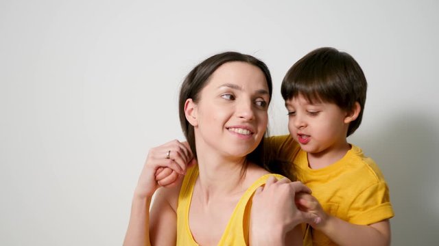 mother with her son a child of three years in yellow t-shirts, stands against a white wall in the studio and wants to kiss her