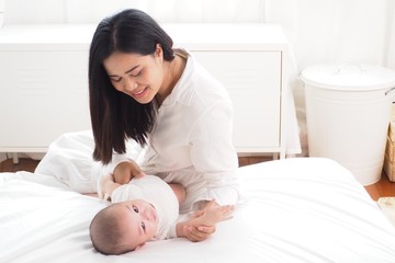 Obraz na płótnie Canvas Beautiful Asian mother looking at and playing with her cute baby lying on bed in the bedroom in white and warm tone. Happy motherhood, unconditional love, family lifestyle concept