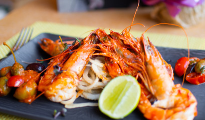 Sauteed shrimps in tomato, capers and olives sauce. Black plate in a restaurant setting background