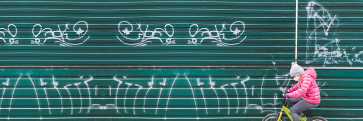 Girl in protective mask and pink jacket riding bike during quarantine. New normal life city. Little lady enjoying bike ride on her way to school or park. Sport for kids and eco transport concept.