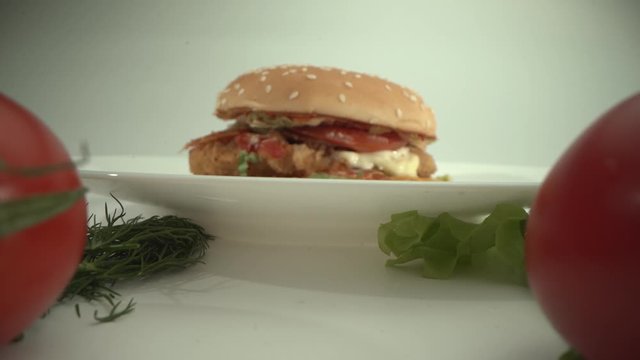 Delicious cheeseburger lies on a plate surrounded by fresh tomatoes, parsley, cheese. White background. Laowa 24 mm