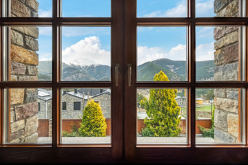Window with a view to the ridge of mountains, snow-capped peak, clouds, sky, trees and houses in Andorra.