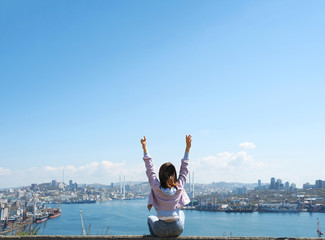  brunette girl in a purple denim jacket and blue jeans raises her hands up and looks at a large modern sea city with a port and cable-stayed bridge. rear view, freedom of movement, end of quarantine