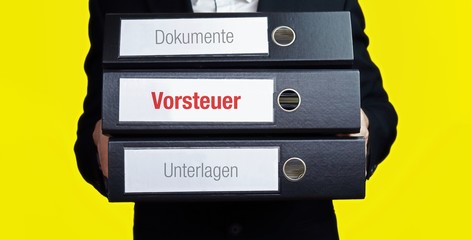 Vorsteuer. Mann im Anzug trägt einen Stapel Aktenordner. Hintergrund gelb. Ein Ordner hat eine rote Beschriftung. Konzept für Business, Statistik, Finanzen © MQ-Illustrations