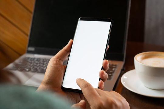 Mockup smartphone image. Woman's hand holding black mobile phone with blank white screen  with coffee cup and laptop on table .