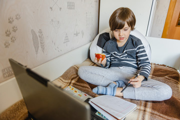 Distance learning for children during the coronavirus epidemic. The boy sits on the bed and receives a school assignment using the Internet and a laptop.