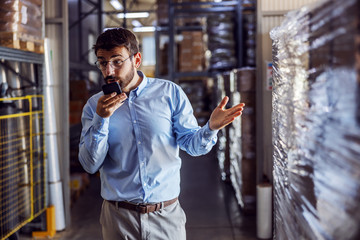 Young bearded businessman having phone conversation with customer while standing in warehouse.