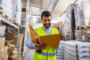 Young smiling employee in vest holding folder with documents and checking out inventory.