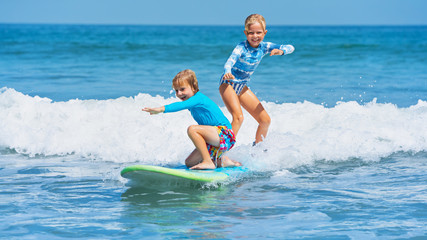 Happy baby boy and girl - young surfers ride with fun on one surfboard. Active family lifestyle, kids outdoor water sport lessons, swimming activity in surf camp. Sea beach summer holiday with child. - obrazy, fototapety, plakaty