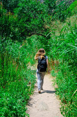 Obraz premium tourist girl with a backpack goes along a path in the mountains among the foliage of trees and grass