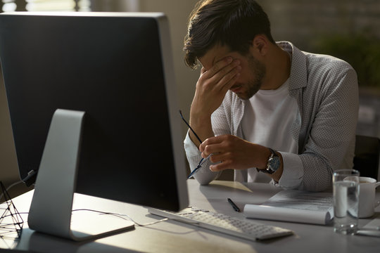 Exhausted businessman holding his head in pain while working on desktop PC in the office. - Powered by Adobe