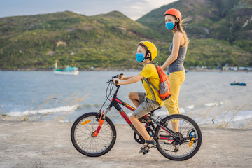 Active school kid boy and his mom in medical mask and safety helmet riding a bike with backpack on sunny day. Happy child biking on way to school. You need to go to school in a mask because of the