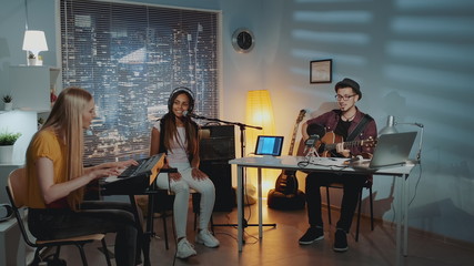 Fototapeta na wymiar Multiracial amateur band is rehearsing by singing playing keyboard and guitar in home studio with cozy interior in the evening