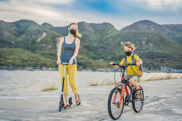 Fototapeta premium Active school kid boy and his mom in medical mask riding a bike with backpack on sunny day. Happy child biking on way to school. You need to go to school in a mask because of the coronavirus epidemic