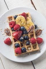 Waffle topped with banana slices, oat, pecans and berries served on a white plate over a white rustic wooden table. Top view.