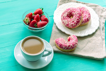 Cup of coffee latte, bowl with fresh strawberries and white plate with pink donuts on turquoise painted wooden table