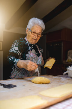 Caucasian grandmother, senior old woman cooking in her kitchen traditional recipes of Italian cuisine. Fresh handmade pasta with eggs and flour, rolling dough on wooden cutting board.