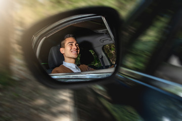 Reflection in car side mirror of a young smiling caucasian man driving. Car in action with blurred background.