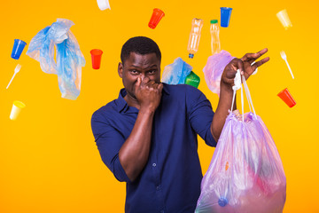 Environmental pollution, plastic recycling problem and waste disposal concept - angry african american man holding garbage bag on yellow background. He is feel smell of trash.
