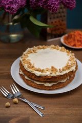 Carrot cake, hazel nuts, grated carrot and bouquet of purple lilac on the wooden table