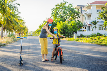Naklejka premium Active school kid boy and his mom in medical mask and safety helmet riding a bike with backpack on sunny day. Happy child biking on way to school. You need to go to school in a mask because of the