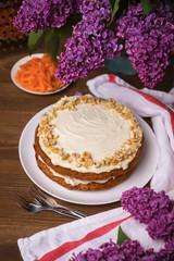 Carrot cake with hazel nuts, grated carrot and bouquet of purple lilac on the wooden table