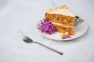 Piece of carrot cake decorated with lilac flowers, and dessert fork on a white tablecloth