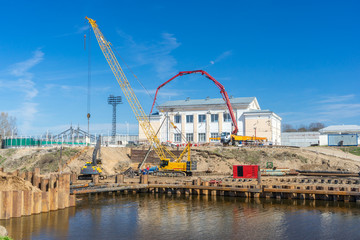 Naklejka premium Construction of pedestrian bridge over river with special heavy machinery and equipment on sunny day. Working process of building supports