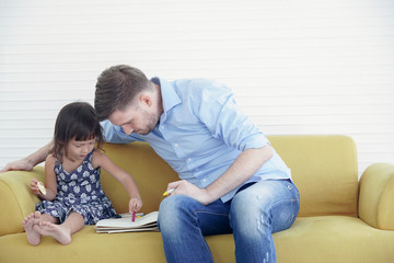 A Caucasian family spend time together, Father looking little daughter while she is drawing and coloring on a book on the yellow sofa at home