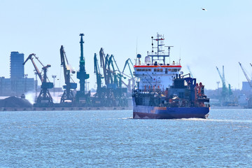 Fototapeta premium View of commercial cargo port with container bridge cranes and a ship entering port on a sunny day.