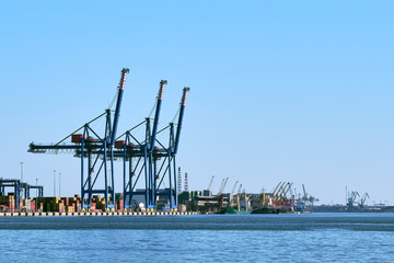 Obraz premium View of port cranes, loading bridges and container terminal on a clear sunny day and cloudless sky. Sea trade, transportation, commerce and cargo
